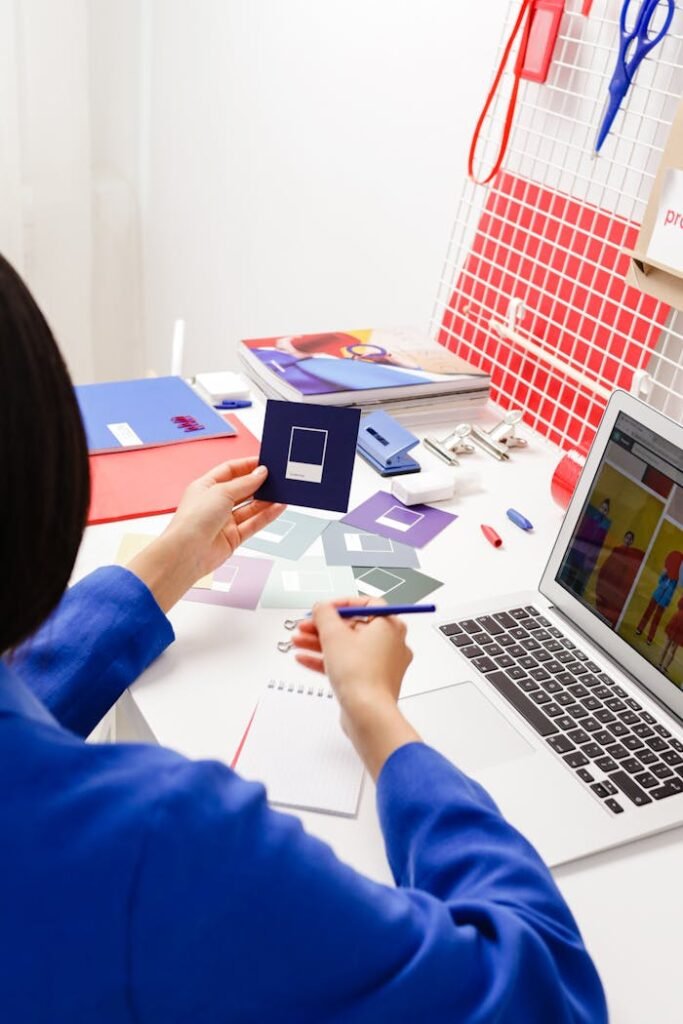 A creative professional in a blue jacket working on design tasks at a desk with a laptop and color swatches.